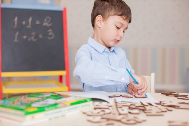 Sevimli küçük çocuk masada oturan ve eğitim mavi tişört giyiyor. Ahşap harfler, sayılar, kitap copybooks ve renkli kalemler ve kalem masada. Duvar ve tahta arka plan bulanık