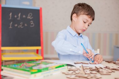 Sevimli küçük çocuk masada oturan ve eğitim mavi tişört giyiyor. Ahşap harfler, sayılar, kitap copybooks ve renkli kalemler ve kalem masada. Duvar ve tahta arka plan bulanık