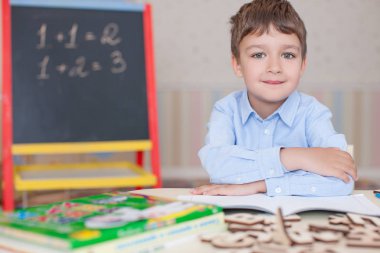 Sevimli küçük çocuk masada oturan ve eğitim mavi tişört giyiyor. Ahşap harfler, sayılar, kitap copybooks ve renkli kalemler ve kalem masada. Duvar ve tahta arka plan bulanık