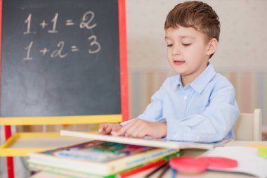 Sevimli küçük çocuk masada oturan ve eğitim mavi tişört giyiyor. Ahşap harfler, sayılar, kitap copybooks ve renkli kalemler ve kalem masada. Duvar ve tahta arka plan bulanık