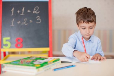 Sevimli küçük çocuk masada oturan ve eğitim mavi tişört giyiyor. Ahşap harfler, sayılar, kitap copybooks ve renkli kalemler ve kalem masada. Duvar ve tahta arka plan bulanık