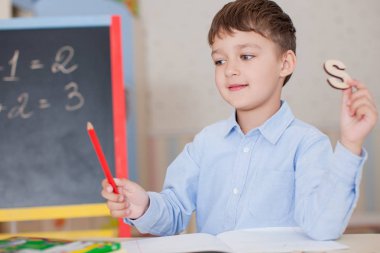 Sevimli küçük çocuk masada oturan ve eğitim mavi tişört giyiyor. Ahşap harfler, sayılar, kitap copybooks ve renkli kalemler ve kalem masada. Duvar ve tahta arka plan bulanık