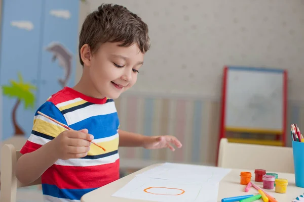 快乐的男孩 穿着五颜六色的 Shirt 在白纸上画画 站在桌子上 用彩色颜料和铅笔 — 图库照片