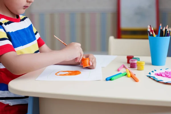 快乐的男孩 穿着五颜六色的 Shirt 在白纸上画画 站在桌子上 用彩色颜料和铅笔 — 图库照片