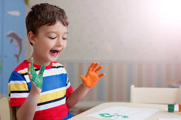 可爱的小男孩穿着五颜六色的条纹衬衫坐在桌边画画 愉快的孩子与绘的手坐在家 模糊的家庭内部背景 — 图库照片