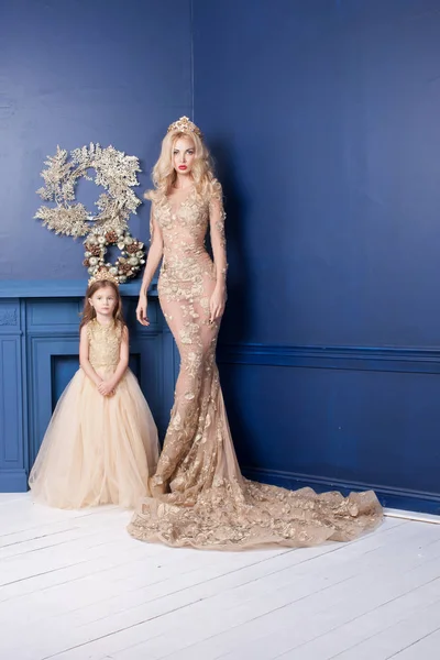 工作室拍摄的美丽的金发女郎与可爱的小女儿穿着金色的节日礼服 愉快的家庭站立在装饰的家庭内部 在蓝色墙壁背景 — 图库照片