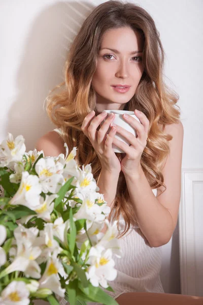 Sun shining into bedroom. Closeup of beauty woman in white pajamas with coffee cup sitting on floor near flowers in bedroom