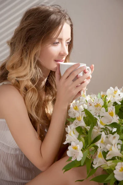 Sun shining into bedroom. Closeup of beauty woman in white pajamas with coffee cup sitting on floor near flowers in bedroom