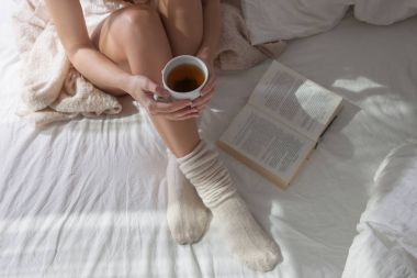 Yatakta güzel kadın bacaklar closeup. Çay içme ve kitap okuma kadın. Yün çorap yatakta üzerinde oturan kız