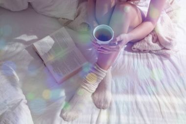 Yatakta güzel kadın bacaklar closeup. Çay içme ve kitap okuma kadın. Yün çorap yatakta üzerinde oturan kız