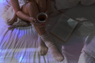 Yatakta güzel kadın bacaklar closeup. Çay içme ve kitap okuma kadın. Yün çorap yatakta üzerinde oturan kız