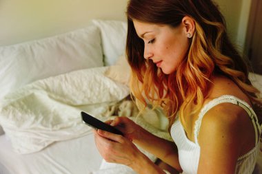 Yatak odasında yatağın üzerinde oturan ve modern smartphone kullanarak güzellik genç kadın