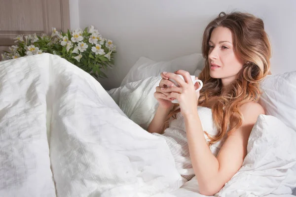 Beautiful young woman wearing white dress lying on bed under white blanket and holding cup of morning coffee