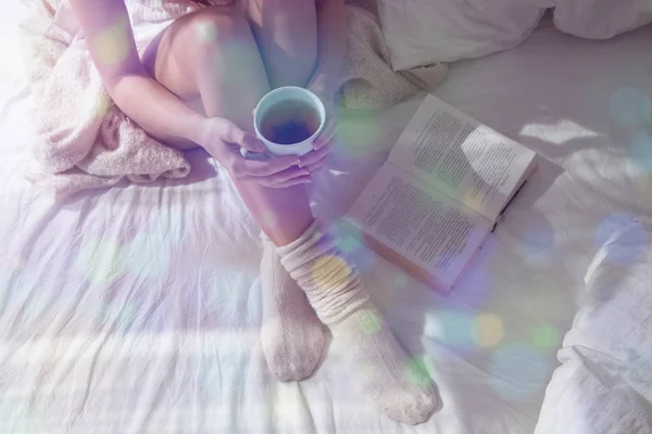 Closeup of beautiful female legs in bed. Woman drinking tea and reading book. Girl sitting on bed in woolen socks