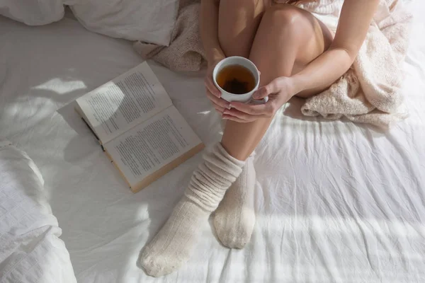 在床上的美丽的女性腿特写 女人喝茶和看书 女孩坐在床上的羊毛袜子 — 图库照片