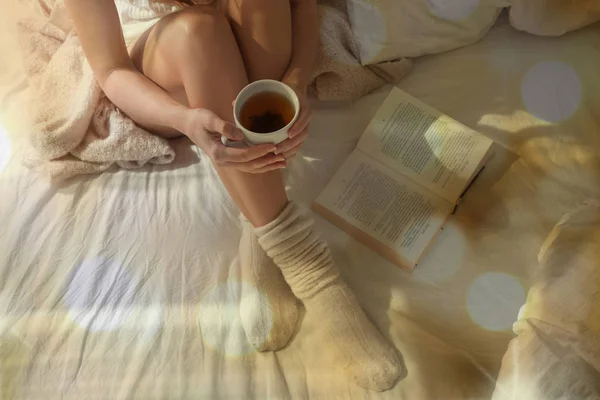 Closeup of beautiful female legs in bed. Woman drinking tea and reading book. Girl sitting on bed in woolen socks