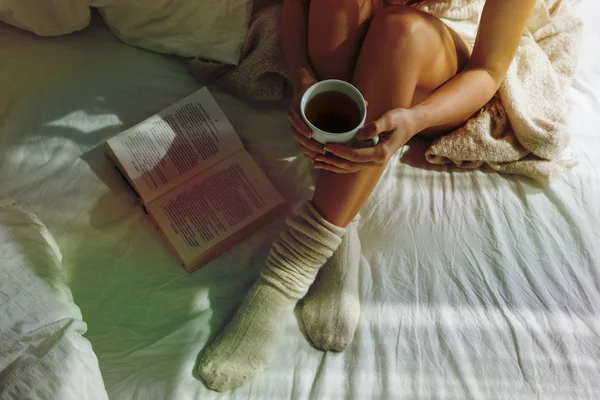 Closeup of beautiful female legs in bed. Woman drinking tea and reading book. Girl sitting on bed in woolen socks