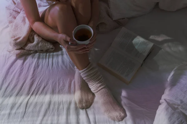 Closeup of beautiful female legs in bed. Woman drinking tea and reading book. Girl sitting on bed in woolen socks