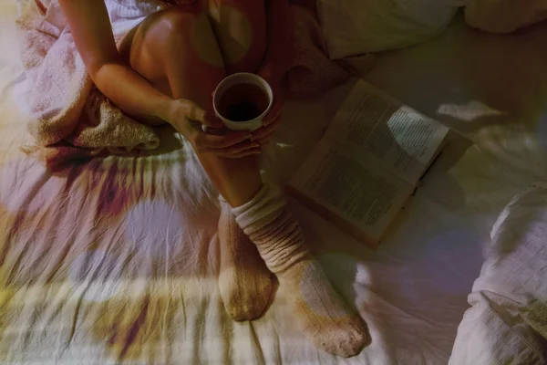 Closeup of beautiful female legs in bed. Woman drinking tea and reading book. Girl sitting on bed in woolen socks