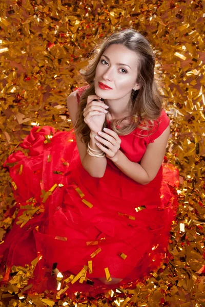 Beautiful young woman with stylish haircut wearing red dress, sitting on golden studio background surrounded by golden confetti, holiday celebration concept