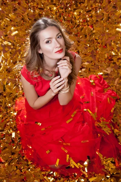 美丽的年轻女子与时尚的发型穿着红色的礼服 坐在金色的五彩纸屑的背景 节日庆典的概念 — 图库照片