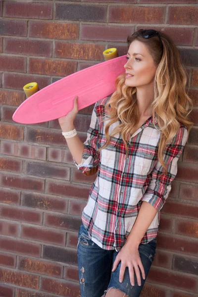 美丽的年轻女子站在砖墙背景与粉红色滑板手 — 图库照片