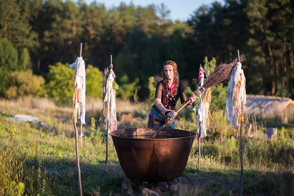 Hermosa bruja preparar una poción en el caldero grande. ¿Qué? Muchos amuletos en las manos de la hechicera. .. Hag agitar el agua con una escoba — Foto de Stock