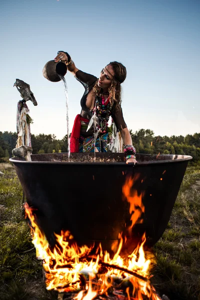 Beautiful witch brew a potion in the big cauldron. ? lot of amulets on enchantress hands. Boiling elixir in the pot. Hag pours water from a jug into a pot — Stock Photo, Image