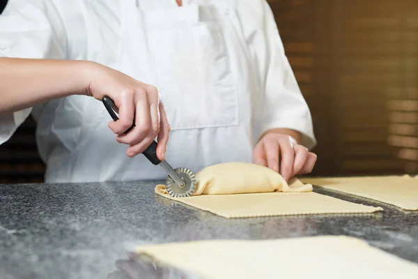 Baker hace algunos rollos de masa cruda y relleno, utilizando un cuchillo especial para la masa — Foto de Stock