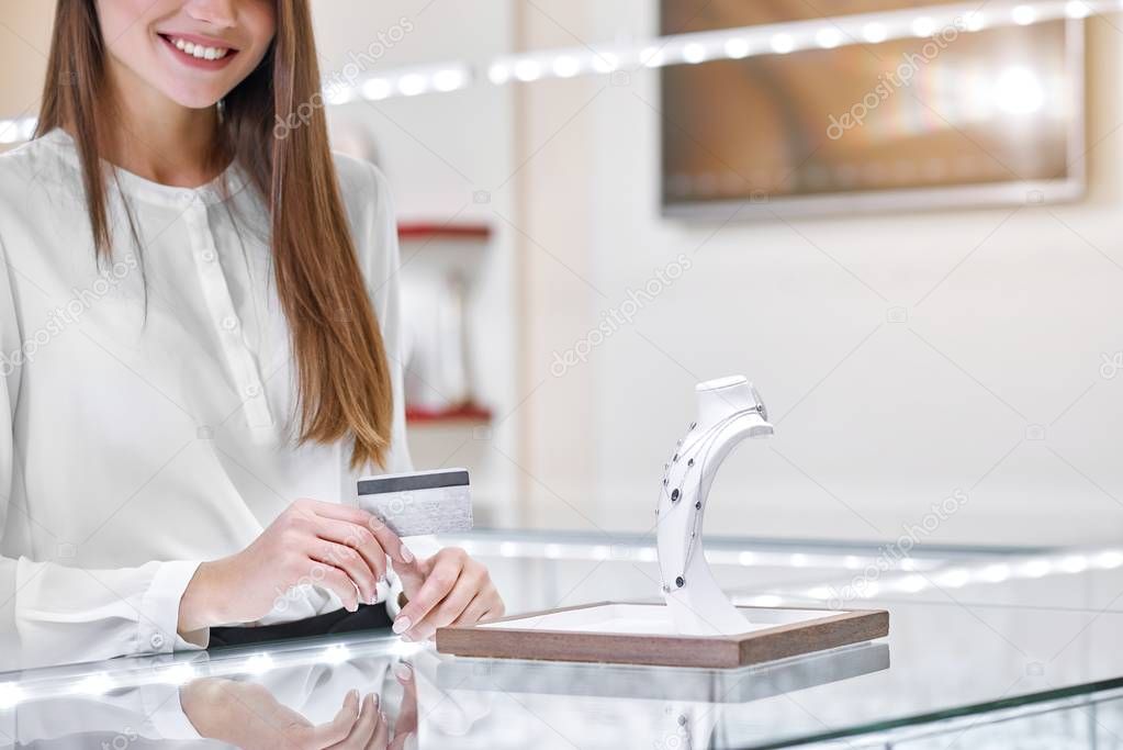Smiling woman near the counter in a jewelry shop is holding a credit card ready to pay for the necklace.