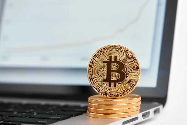 Stack of golden bitcoins with one bitcoin on its edge standing on laptop with financial chart on its screen — Stock Photo, Image