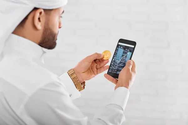 Young Muslim trader keeps golden bitcoin and mobile phone — Stock Photo, Image