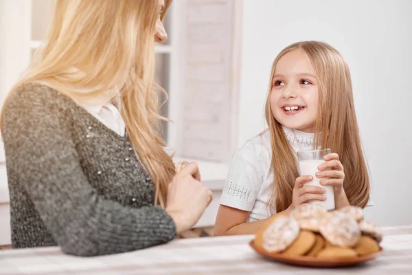 Sorrindo pequena filha senta-se cochilando copo de leite perto da mãe . — Fotografia de Stock