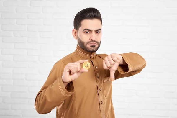 Przystojny młody mężczyzna muzułmańskich jest smutny i zdenerwowany trzymając złote bitcoin i pokazuje kciuk w dół — Zdjęcie stockowe