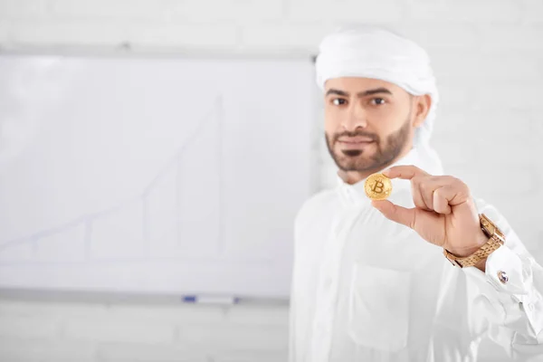 Handsome muslim male model in traditional Islamic clothing holding golden bitcoin in front of white board wall — Stock Photo, Image