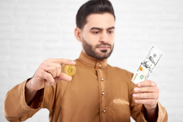 穆斯林男性模式在传统的伊斯兰服装看着百美元, 同时持有黄金比特币在他手中 — 图库照片