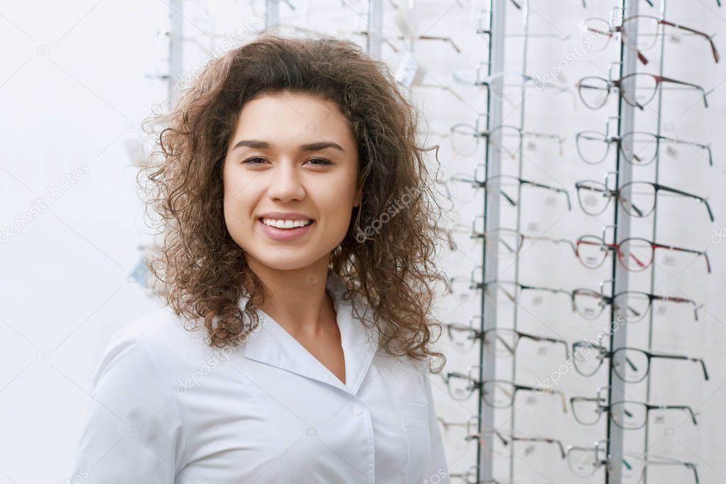 Smiling curly ophthalmologist posing near stand with eyeglasses.