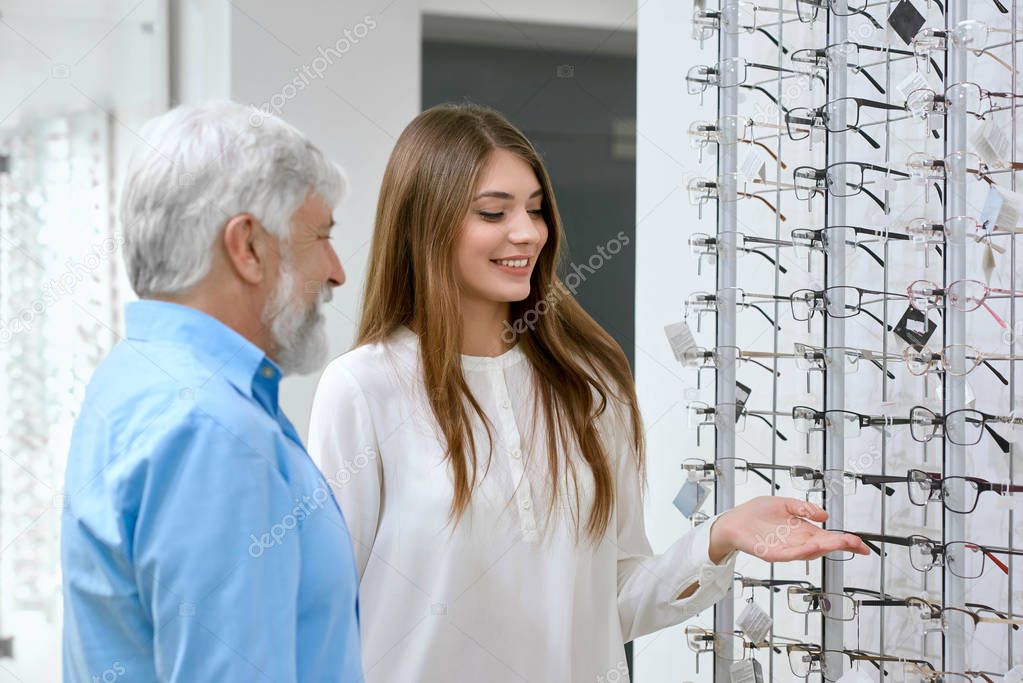 Sideview of pretty smiling assistant showing eyeglasses to old patient.