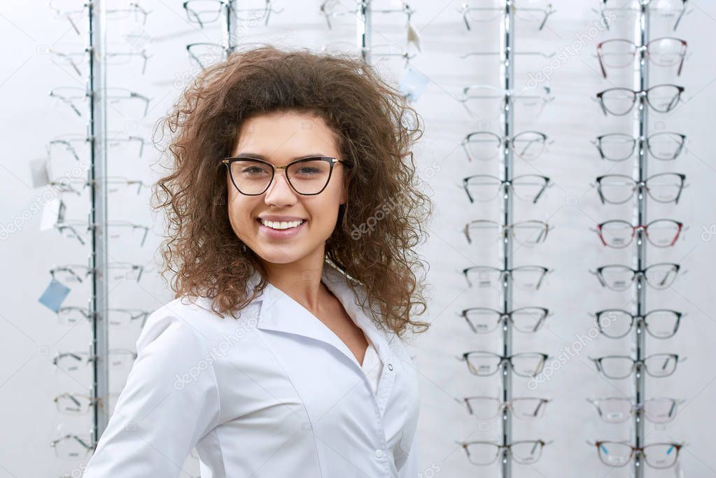 frontview of smiling curly ophthalmologist posing near stand with eyeglasses.