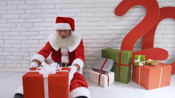 Alter Mann im Weihnachtsmannkostüm sitzt neben Wand mit Geschenkschachteln — Stockvideo