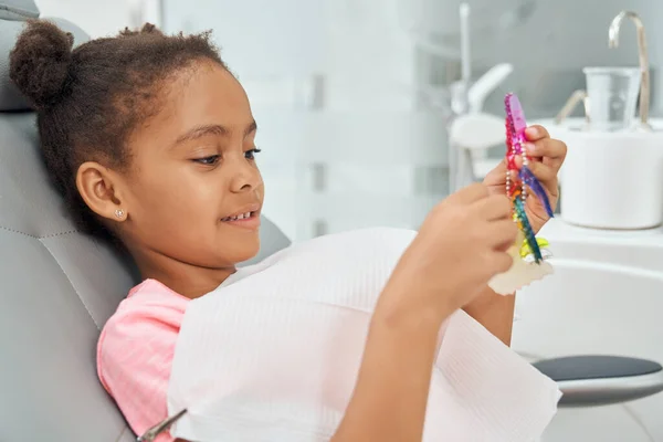 Afrikanisches Kind sitzt im Zahnarztstuhl und hält Zahnspange. — Stockfoto