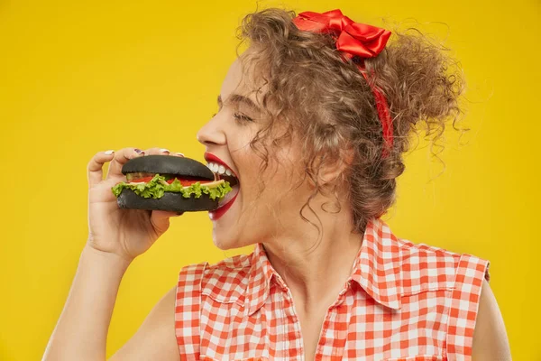 Seitenansicht eines glücklichen jungen Mädchens, das versucht, schwarzen Burger zu essen — Stockfoto