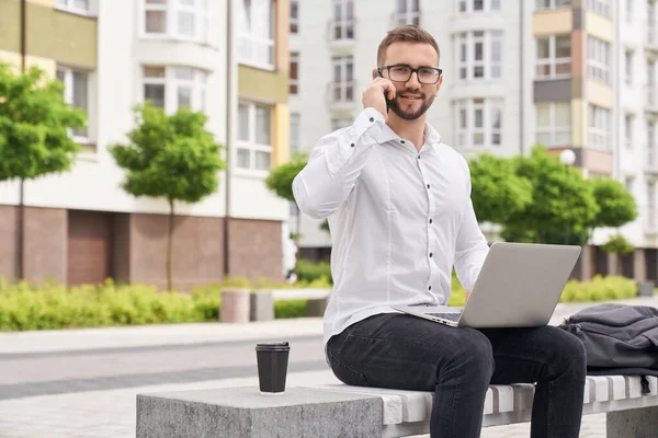 Freelancer com laptop sentado no banco falando por telefone . — Fotografia de Stock