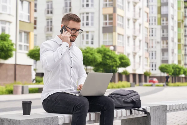 Programador sentado no banco com laptop falando por telefone . — Fotografia de Stock