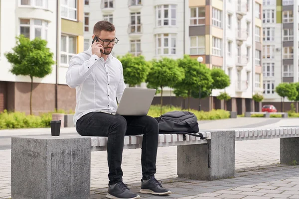 Hombre sentado en el banco, trabajando en el portátil, hablando por teléfono . — Foto de Stock