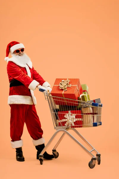 Święty Mikołaj w okularach przeciwsłonecznych z wózkiem wypełnionym prezentami — Zdjęcie stockowe