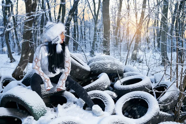 Дівчина в капелюсі сидить на автомобільних шинах в лісі взимку . — стокове фото