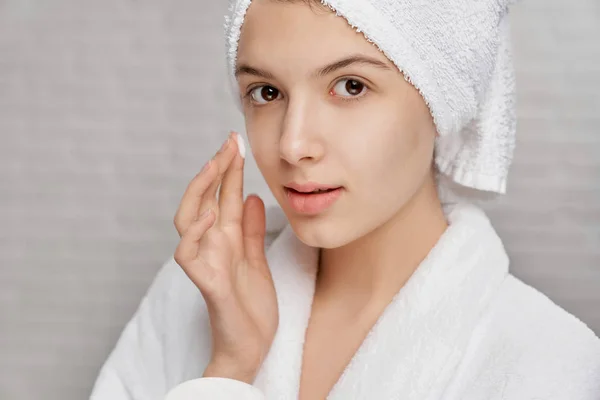 Young woman looking at camera while applying cream on face — Stock Photo, Image