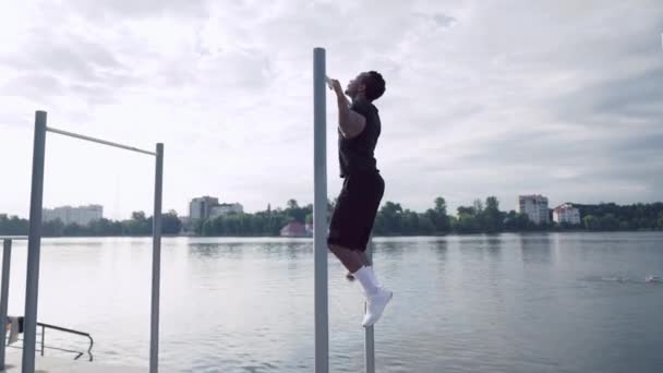 Afro man doing pull ups on horizontal bar near lake — Stockvideo