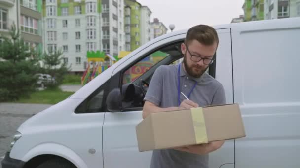 Mladý kurýr stojící poblíž auta se schránkou a balíkem — Stock video